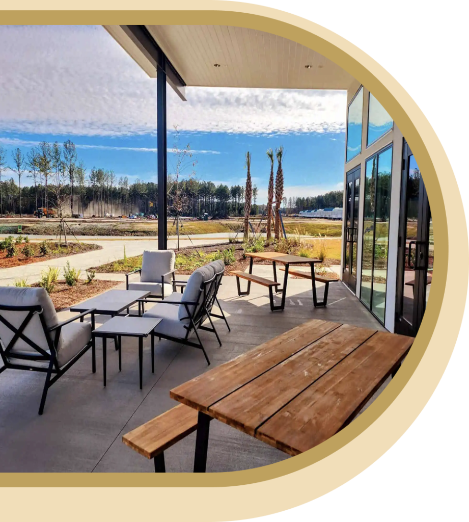 Outdoor patio with wooden picnic tables, cushioned chairs, and a scenic view of landscaped grounds, trees, and a partly cloudy sky.
