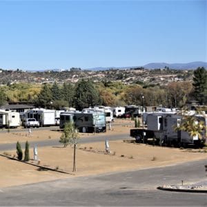 A large RV park with several motorhomes and trailers parked in designated spots, resembling a gallery of modern drive-in homes. The landscape in the background features a mix of trees, open land, and distant hills under a clear blue sky. The area appears to be dry, with sparse vegetation between spaces.