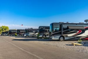 A row of RVs parked at a campsite on a sunny day gives off a cozy, home-like vibe. The sky is clear and blue, with an American flag waving in the background. The ground is covered with gravel, and small green shrubs line the parking area.