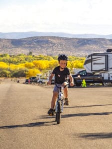 A young child wearing a helmet rides a bike on a paved road with hills and trees in the background, resembling an autumn gallery. An RV is parked to the side, and the trees have bright yellow foliage, indicating fall. A distant figure is walking along the road.