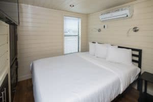 A cozy, modern bedroom with light wooden walls and a large window with blinds exhibits a minimalist charm. The double bed features white bedding and two pillows. Built-in storage, an air conditioning unit above the bed, and two adjustable reading lights add to the gallery-like elegance of the room.