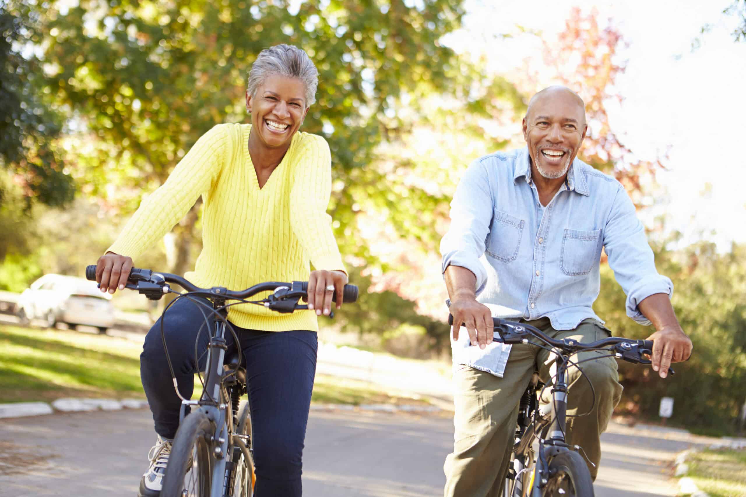 A smiling older couple, embodying the classic snowbirds lifestyle, rides bicycles outdoors on a sunny day. The woman wears a yellow sweater, and the man wears a light blue shirt. They appear happy, riding side by side on a tree-lined path with green and autumn-colored foliage in the background.