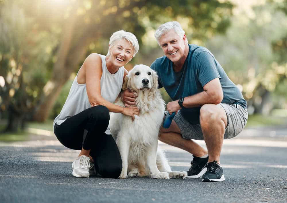 A smiling older couple, seasonal travelers, kneeling on a paved path in a park and posing with a fluffy white dog. The woman has short gray hair and is wearing a white sleeveless top and black leggings. The man has gray hair, a blue T-shirt, and gray shorts. It's a sunny day.