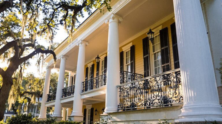 A stately antebellum-inspired mansion with tall white columns and a wraparound porch under oak trees draped in Spanish moss, captured in warm daylight, highlighting the elegant southern architecture and serene ambiance.