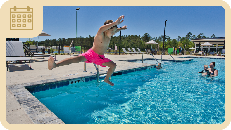 A child in pink swim trunks jumps into an outdoor swimming pool, while two people and a child are in the water nearby under a clear blue sky.