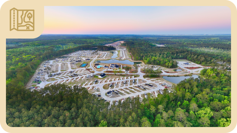 Aerial view of a large, organized campground surrounded by dense forest, with RVs, parked vehicles, winding roads, and ponds visible.
