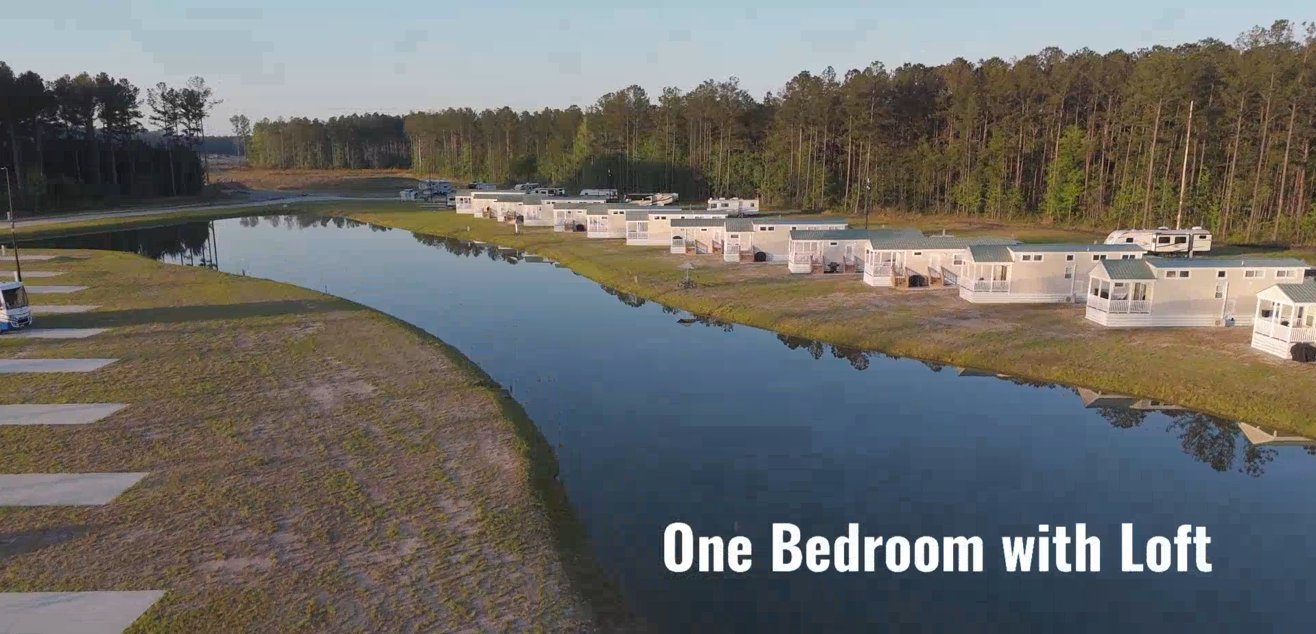 A row of white modular homes is lined up beside a calm river, bordered by trees, with the text “One Bedroom with Loft” in the bottom right corner.