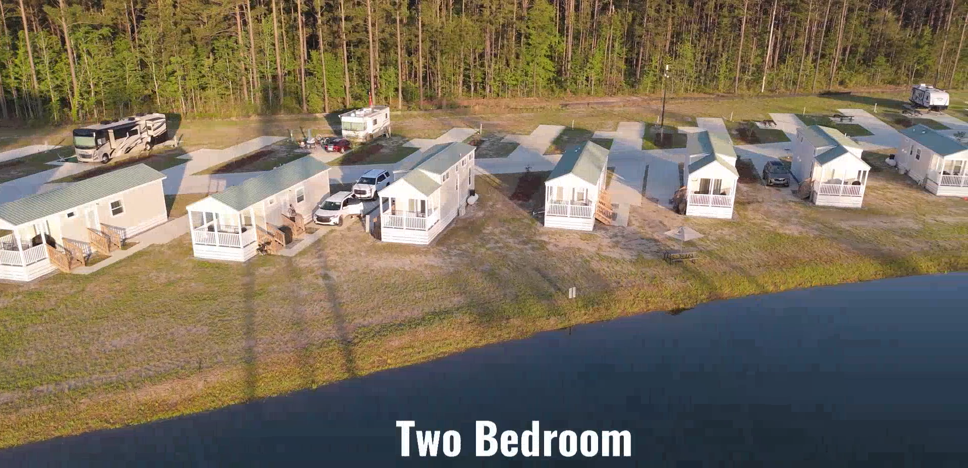A row of two-bedroom modular homes with porches is situated beside a body of water, with parked cars and RVs visible, near a forested area.