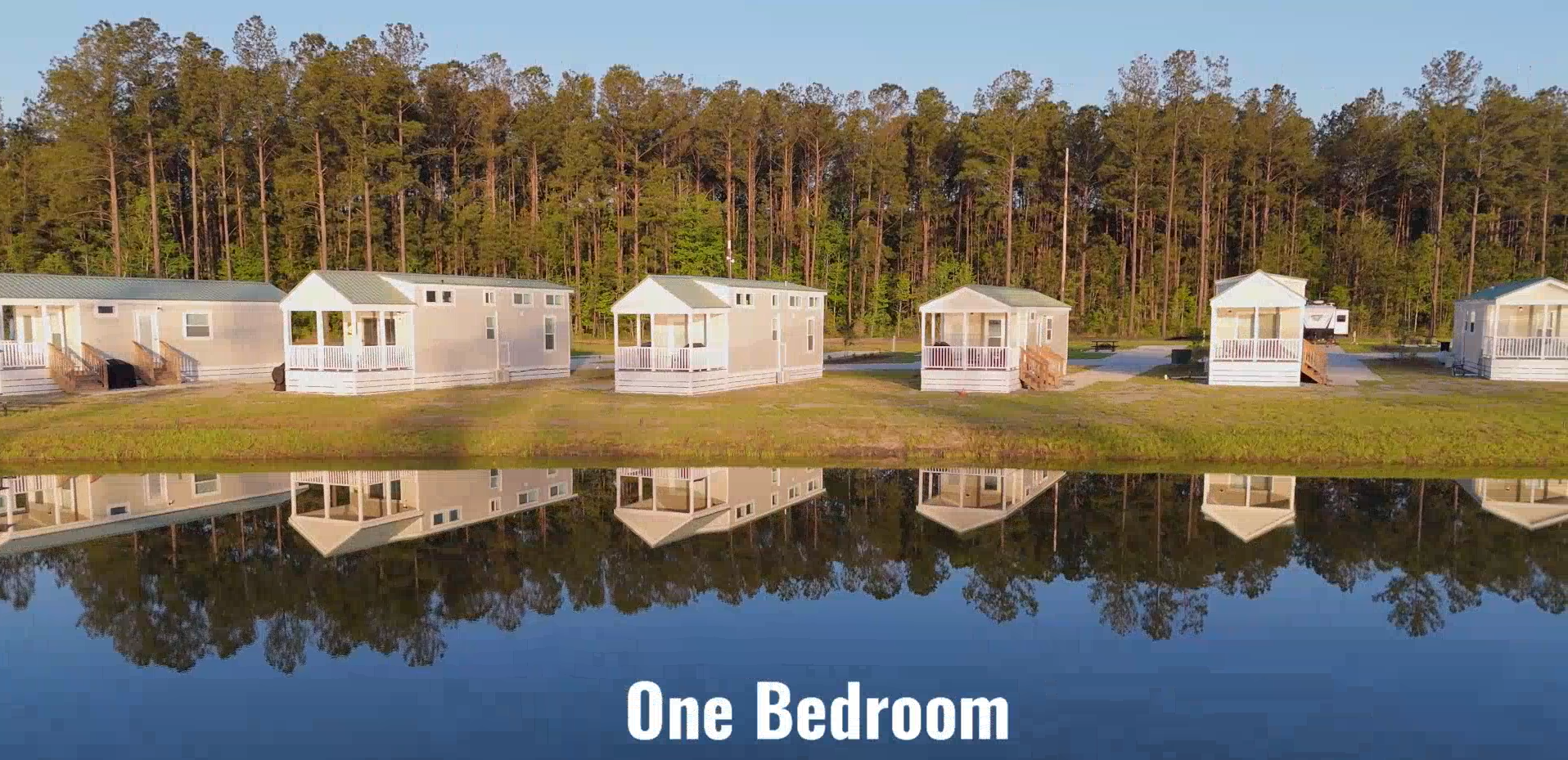 Four small white cabins are lined up beside a pond, reflecting in the water, with a forest of tall trees in the background. Text at the bottom reads "One Bedroom.