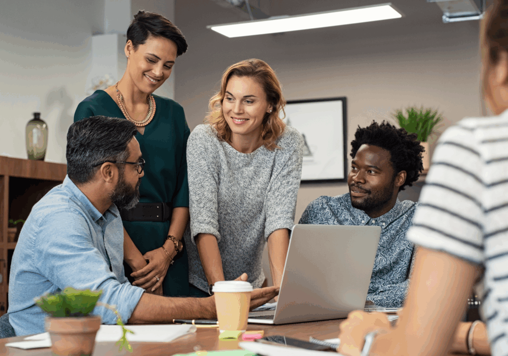 Four people are gathered around a laptop in an office, engaged in a discussion. One person is seated at the laptop, while three others stand or sit nearby, looking at each other and smiling.