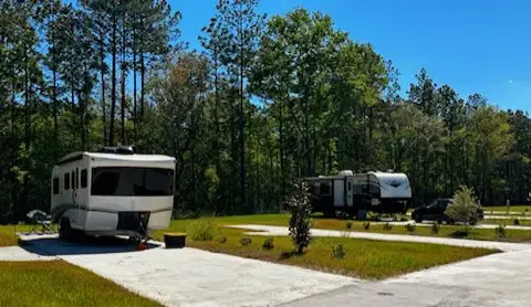 Two RVs are parked on paved pads in a campground surrounded by trees and greenery under a clear blue sky.