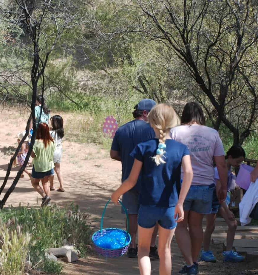 A group of people walk along a dirt path outdoors; a girl in front carries a blue Easter basket. Sparse trees and dry grass surround the area.