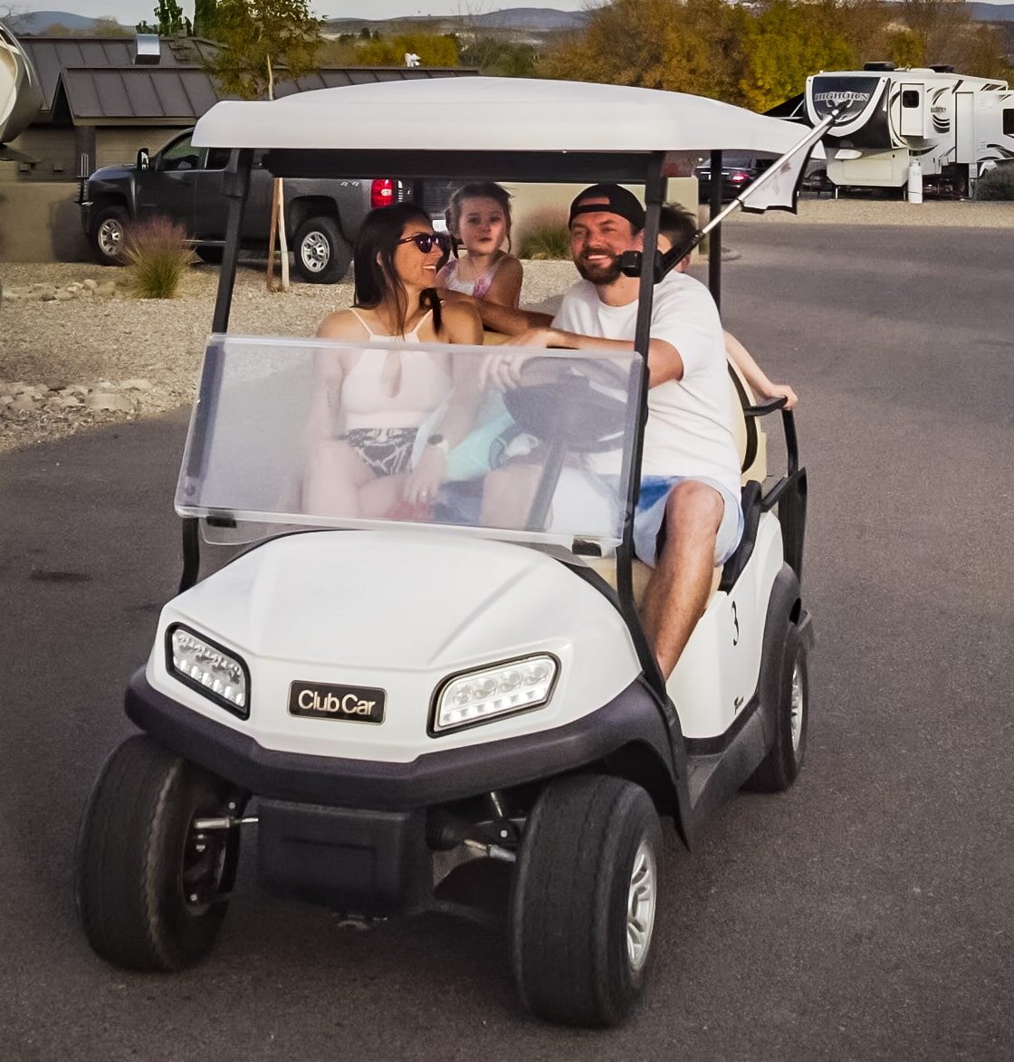 A man and woman sit in the front seats of a white golf cart with two children in the back, driving on a paved road in a campground area.