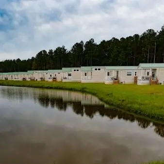 A row of white mobile homes is lined up beside a pond with a grassy area and trees in the background under a cloudy sky.