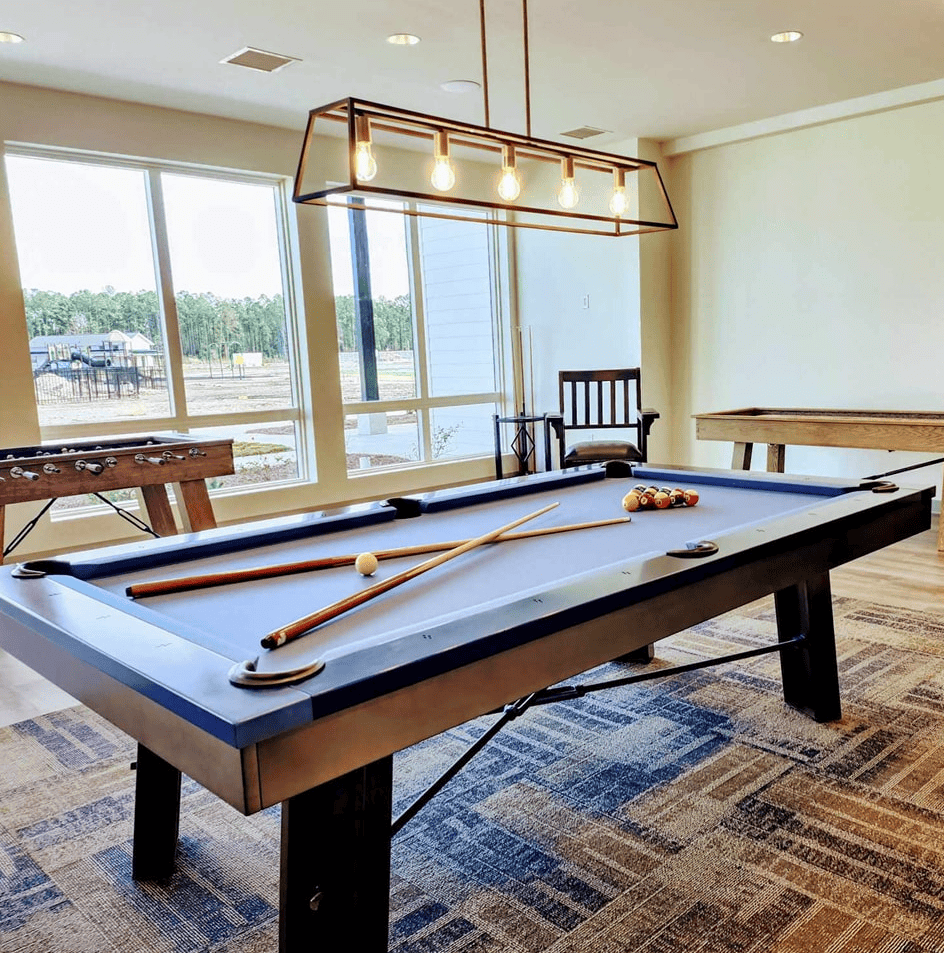 A blue pool table with cues and balls stands in a bright room with large windows; a shuffleboard table and chair are in the background.