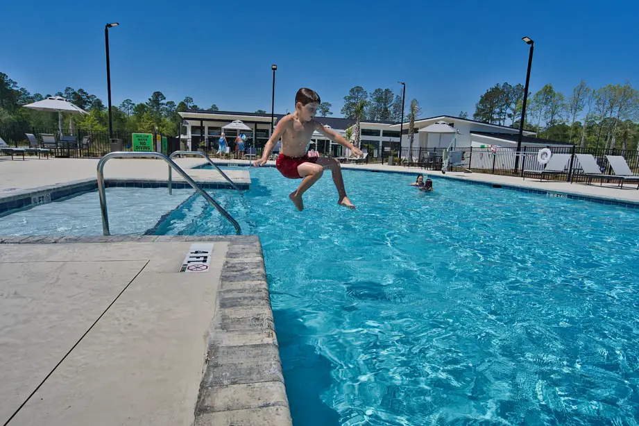 A boy in red swim trunks jumps into a blue outdoor swimming pool on a sunny day; other people relax in the background near the pool.