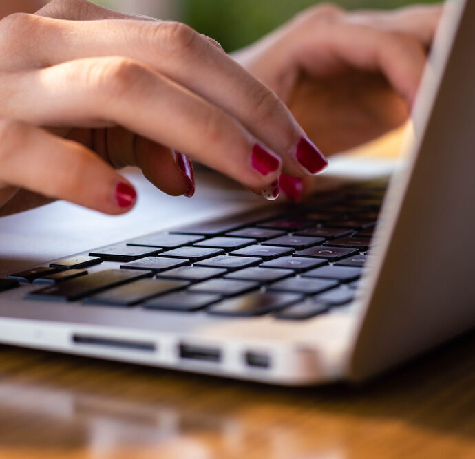 Close-up of hands with red nail polish typing on a laptop keyboard, with the laptop placed on a wooden surface.