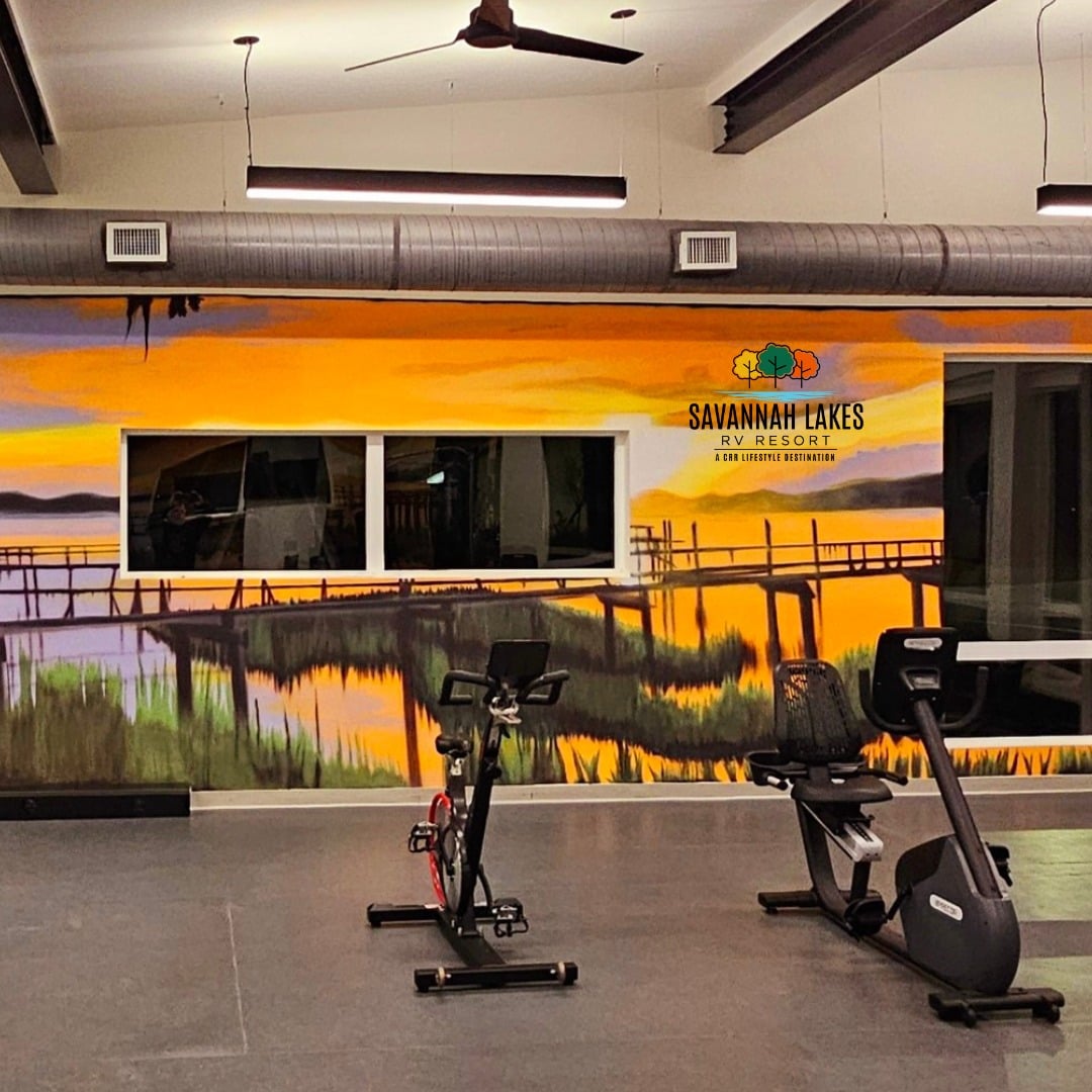 A fitness room with exercise bikes, featuring a wall mural of a lakeside sunset and a "Savannah Lakes RV Resort" sign.