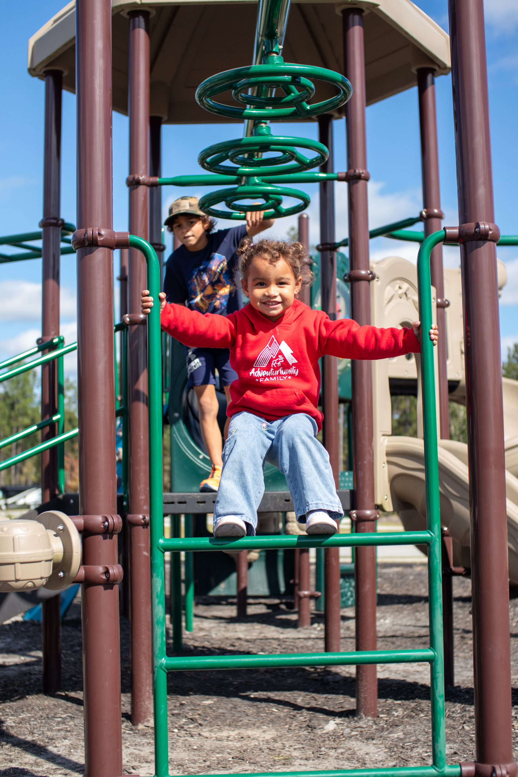 Two children play on a jungle gym at a playground. One child in a red sweatshirt is climbing, while another child stands on a platform in the background.