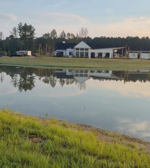 A modern white house with large windows sits near a pond, with its reflection visible in the water; trees and a grassy bank are in the foreground.