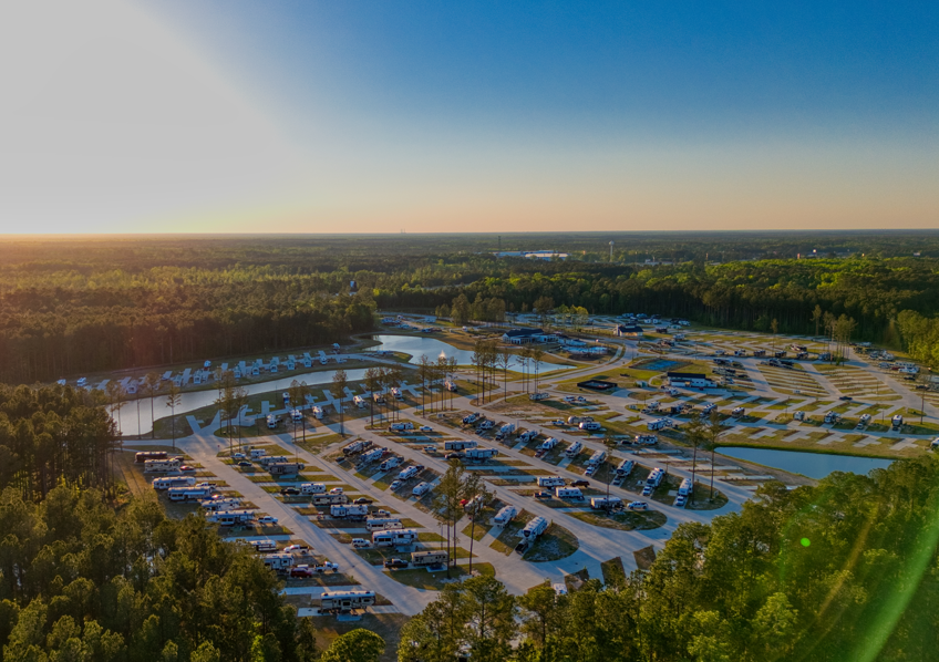 Aerial view of a large RV park with numerous RVs parked in rows, surrounded by trees and small ponds, under a clear sky at sunset.