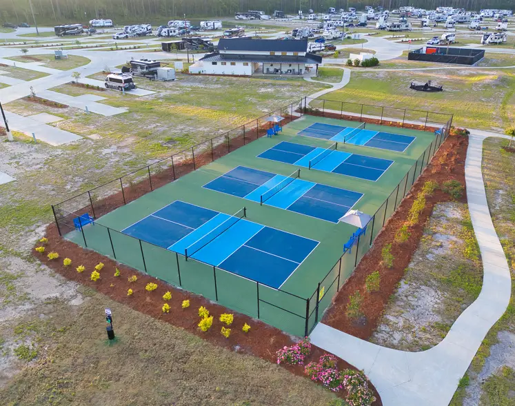 Aerial view of six blue and green pickleball courts surrounded by fencing, walkways, and landscaping, with RVs and a building in the background.