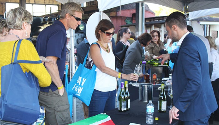 A man in a suit pours wine for a woman at an outdoor event booth while others watch; wine bottles and glasses are on the table.