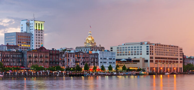 Savannah, Georgia’s riverfront with historic buildings, modern structures, and the gold-domed city hall at sunset, reflected in the water.