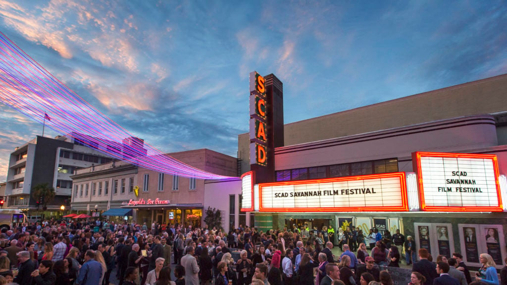 A large crowd gathers outside a theater with a marquee reading "SCAD Savannah Film Festival" at dusk, with colorful lights strung overhead.