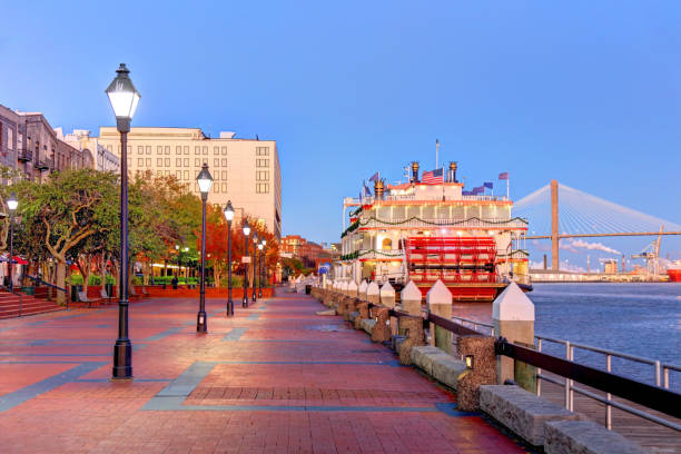 A riverfront walkway with street lamps, trees, and a riverboat docked beside the water; a bridge is visible in the background at sunset.