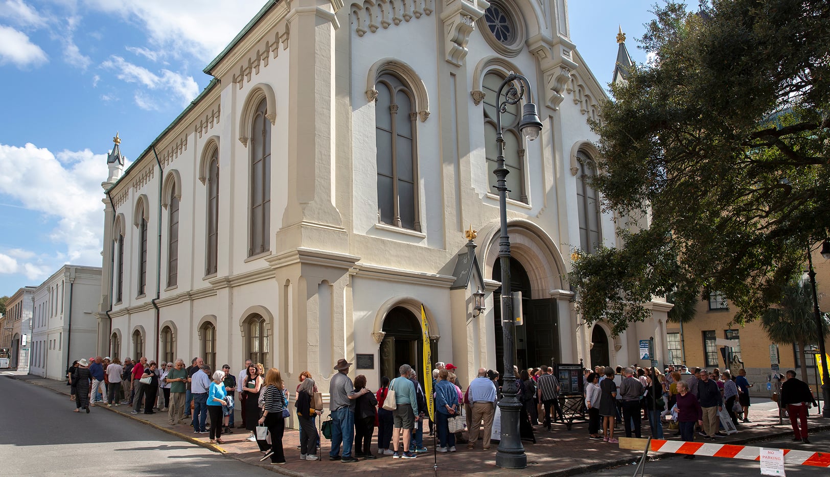 A large group of people stands in line outside the entrance of a white historic building on a sunny day.