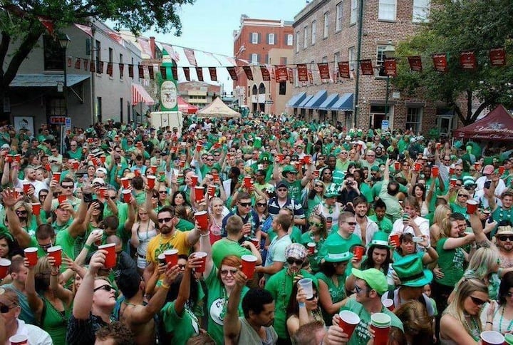 A large crowd of people dressed in green hold up red cups at an outdoor event, likely a St. Patrick's Day celebration, with brick buildings and banners in the background.