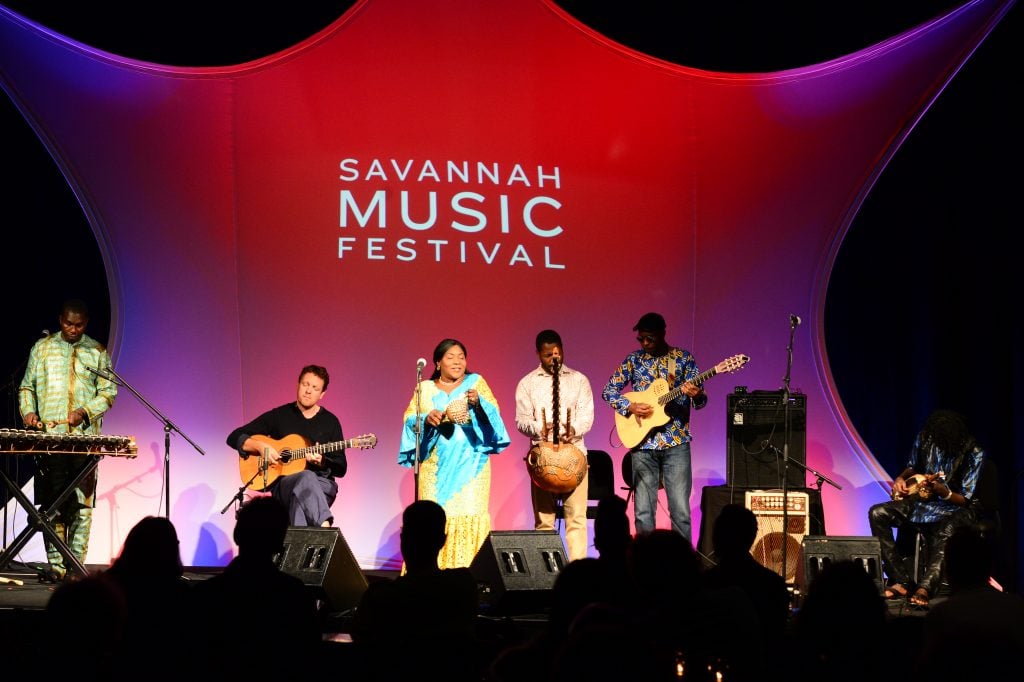 Six musicians perform on stage under red lighting at the Savannah Music Festival, with instruments including guitar, drums, and balafon. Audience silhouettes are visible in the foreground.