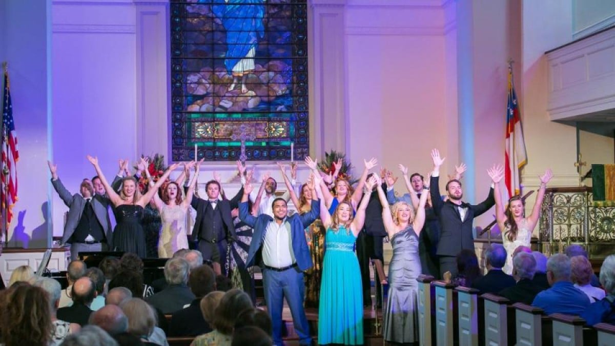 A group of people in formal attire stand at the front of a church, raising their arms while facing an audience, with a stained glass window in the background.