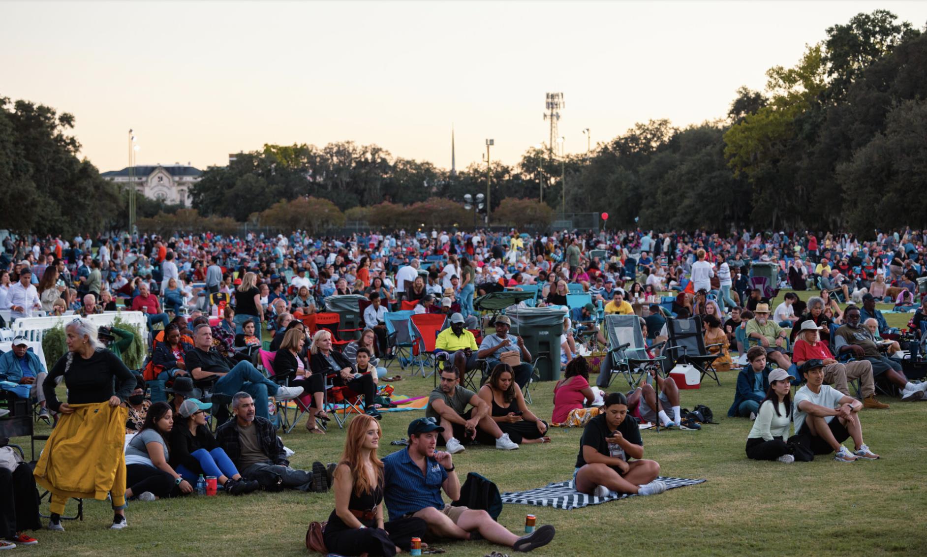 A large crowd of people sits on blankets and lawn chairs in an open grassy area, watching an outdoor event during daylight.