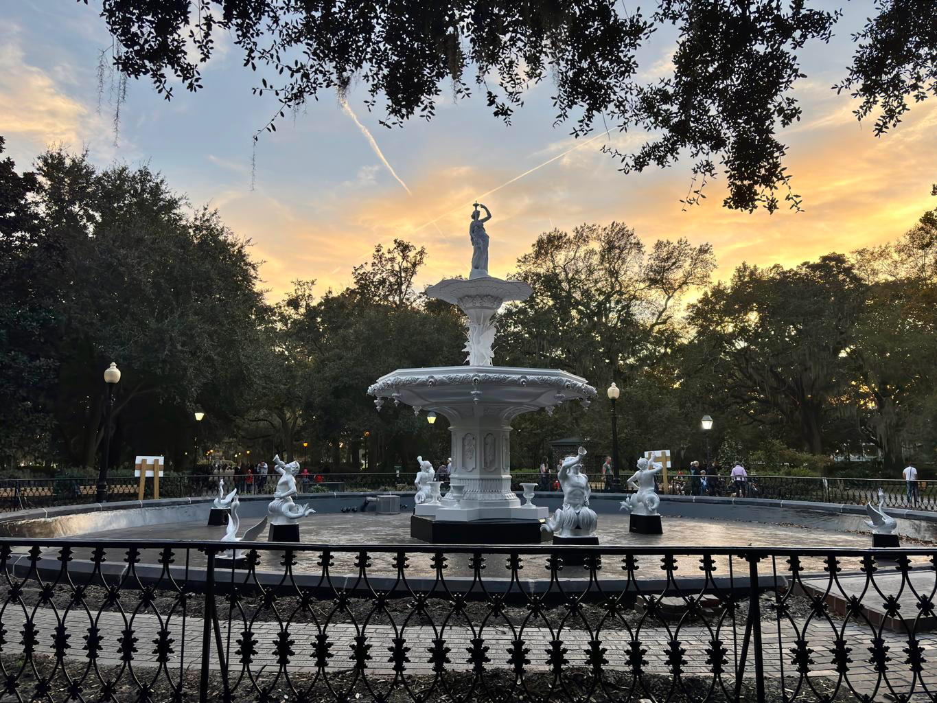 A large white fountain with sculpted figures stands in the center of a park, surrounded by trees and an iron fence at sunset.