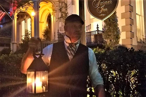 A man dressed in historic clothing holds a lit lantern outside a building with a sign that reads "Eigner Inn.