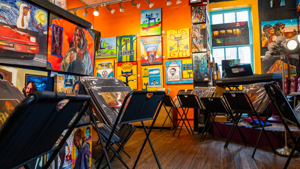 Art gallery room with colorful paintings on bright orange and yellow walls, folding chairs set up, and a wooden floor. Natural and artificial lighting illuminate the space.