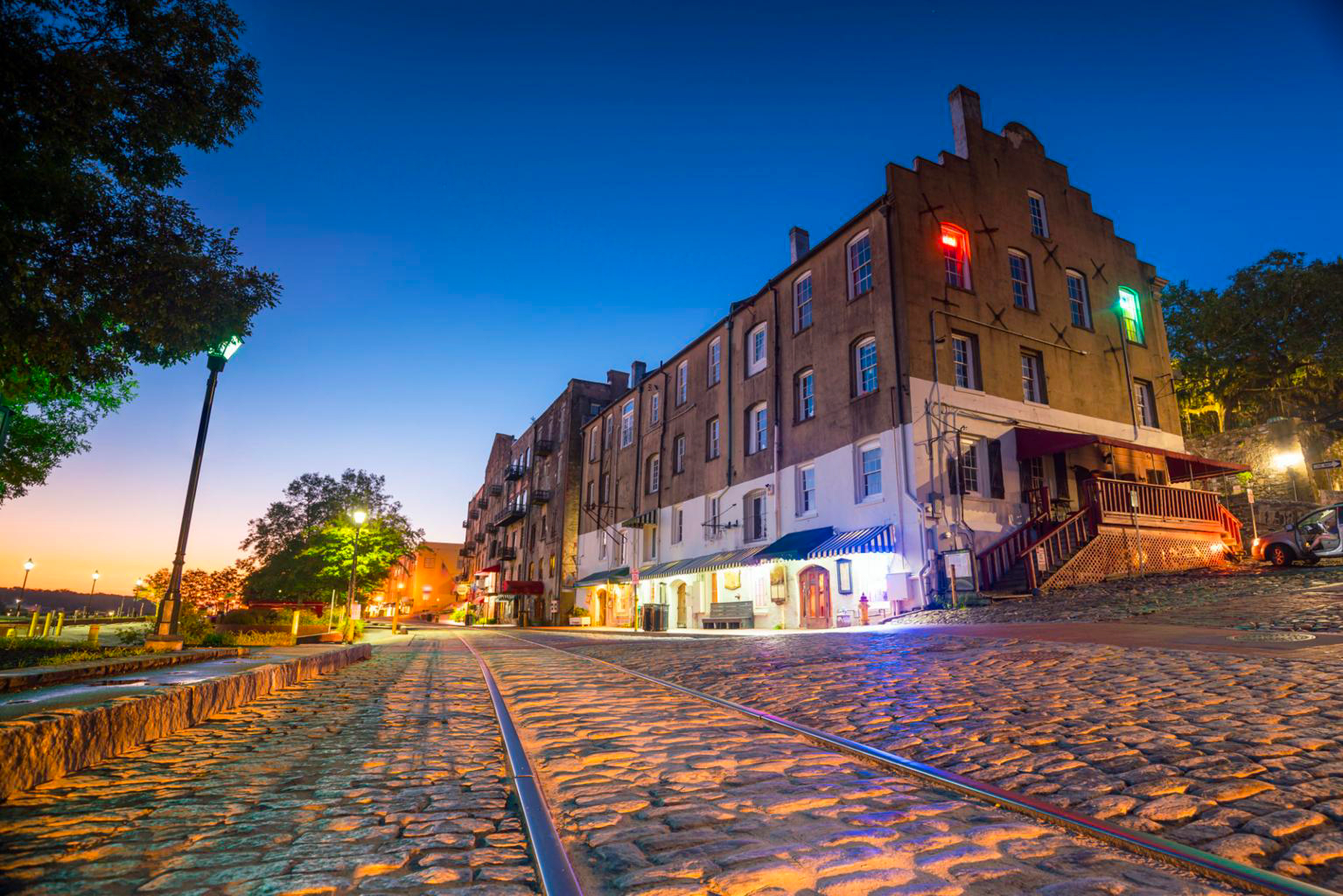Old brick buildings and cobblestone street with train tracks at dusk, illuminated by colorful lights, in a historic city area.