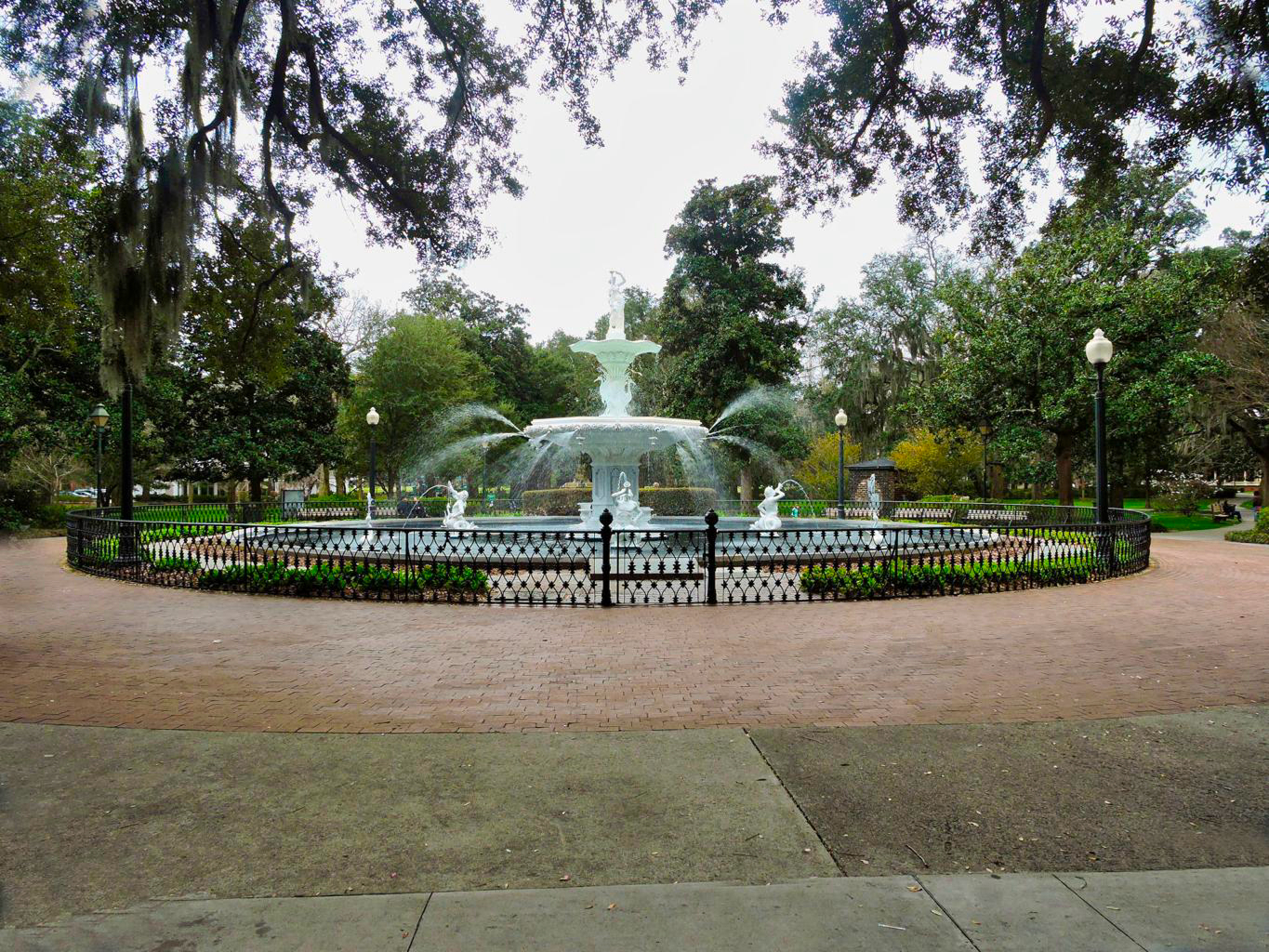 Large white fountain spraying water in the center of a fenced area, surrounded by trees, greenery, and lamp posts in a public park.