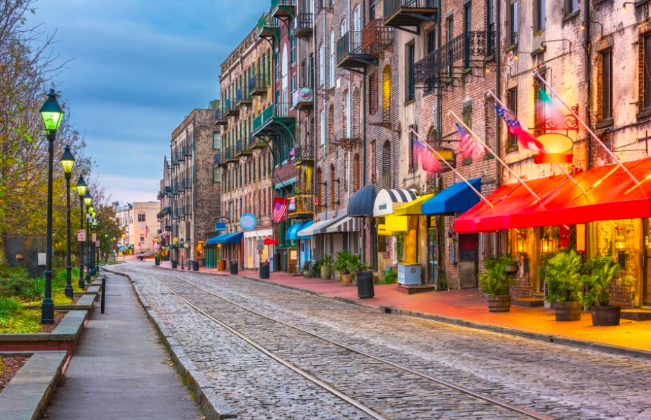 A cobblestone street lined with historic brick buildings, restaurants, shops, and flags, with street lamps and railroad tracks running through the scene.
