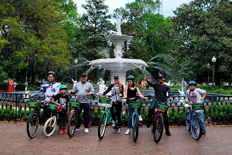 A group of seven people with bicycles pose in front of a large white fountain in a park, surrounded by greenery.