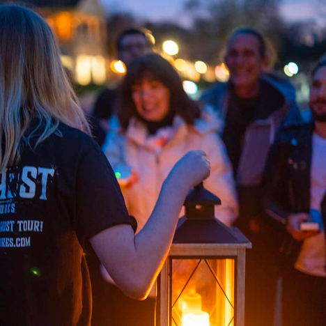 A person holds a lantern in the foreground while a group of people stand and smile in the background at dusk.
