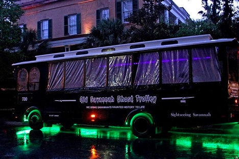 A black Old Savannah Ghost Trolley is parked on a wet street at night, illuminated by green lights underneath and surrounded by historic buildings and trees.
