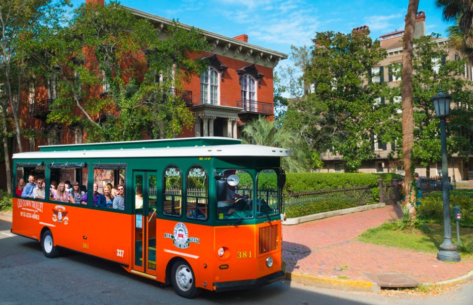 An orange and green trolley with passengers drives past historic brick buildings and trees on a sunny day.