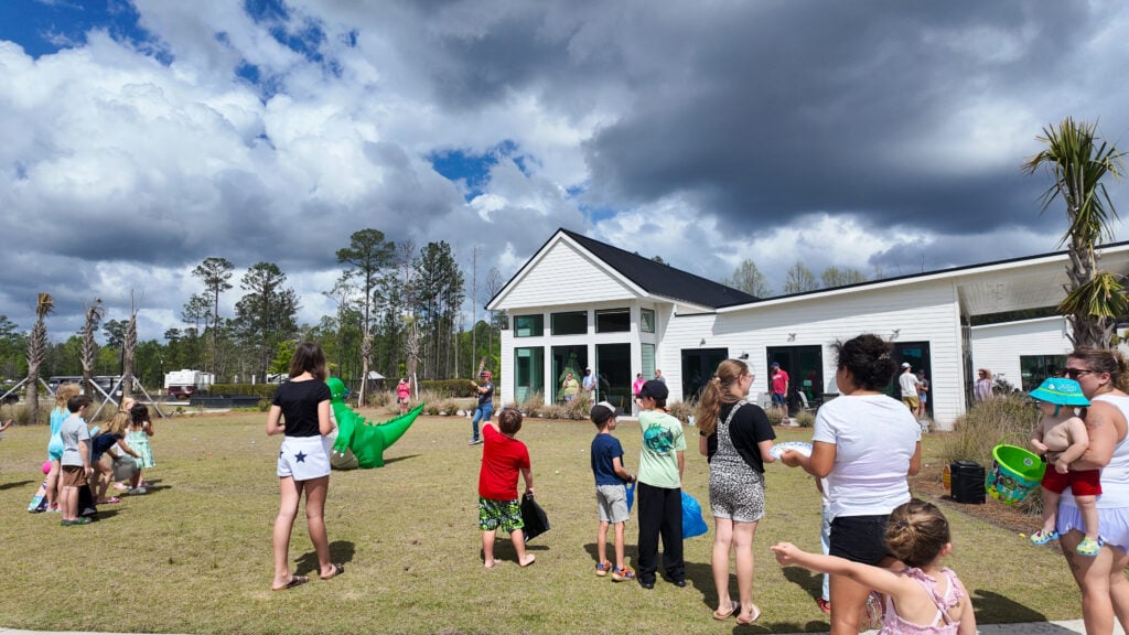 A group of children and adults gather outside a white building for a community event under a partly cloudy sky. Some hold toys and prizes, while others watch activities on the grass.