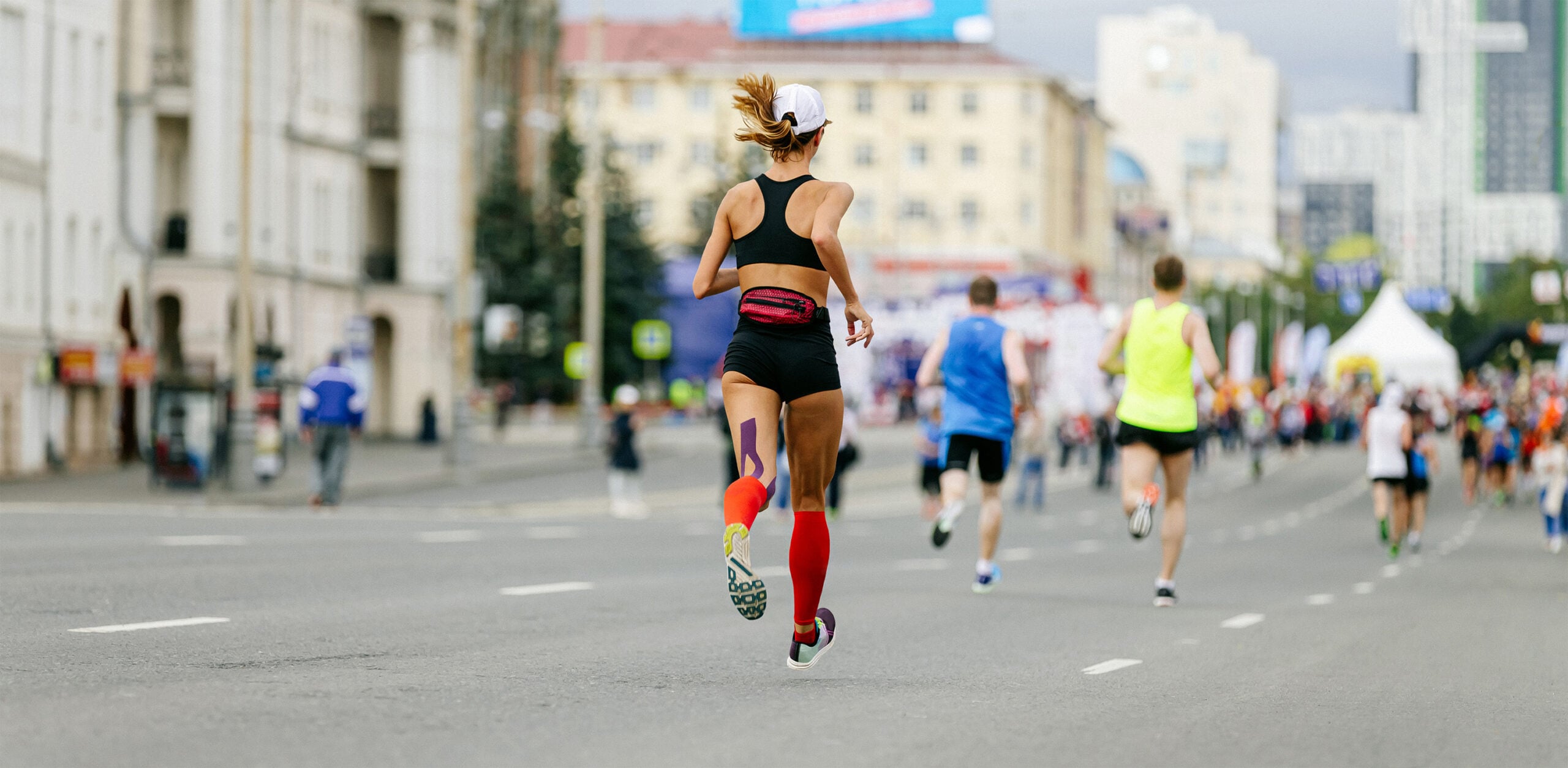 A group of runners participates in a city road race, with a female runner in the foreground wearing athletic gear and a numbered bib.