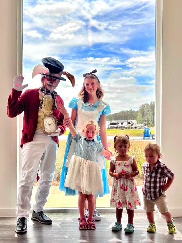 Two adults in "Alice in Wonderland" costumes and three young children pose in front of a large window with a scenic outdoor view.