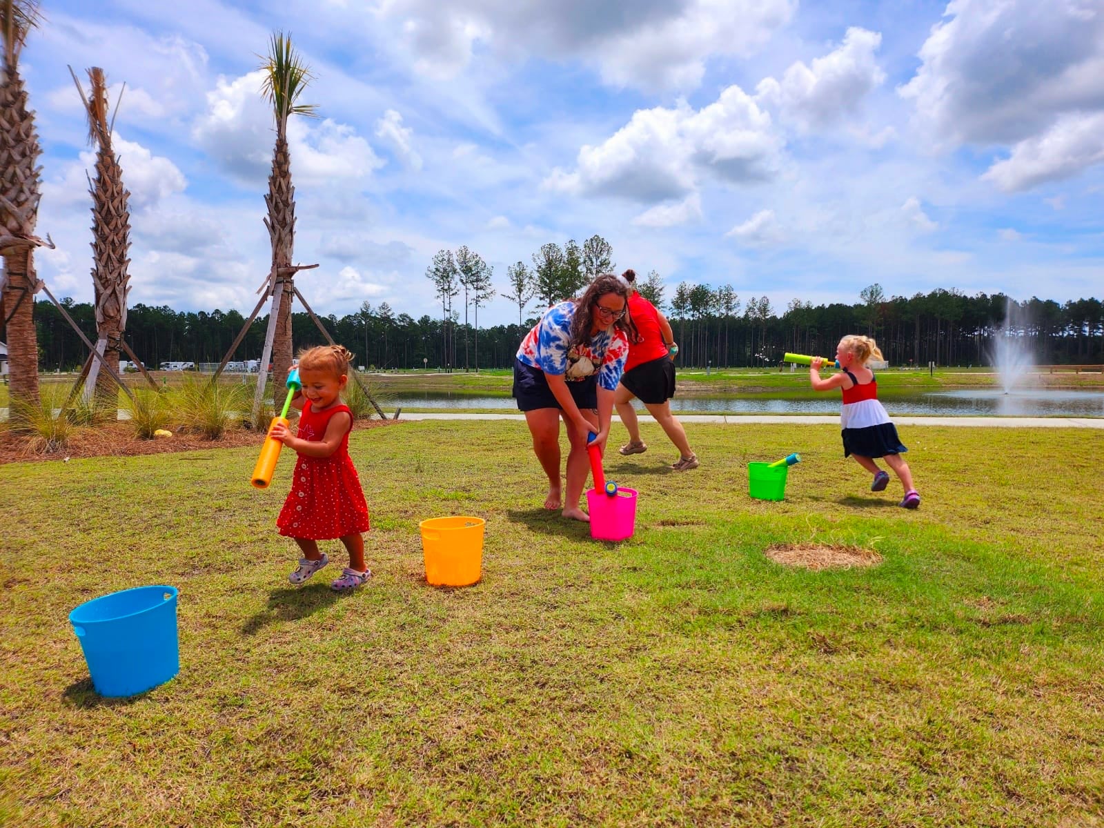 Two women and three young girls play with colorful buckets and tubes on a grassy field near a pond with palm trees and a fountain in the background.