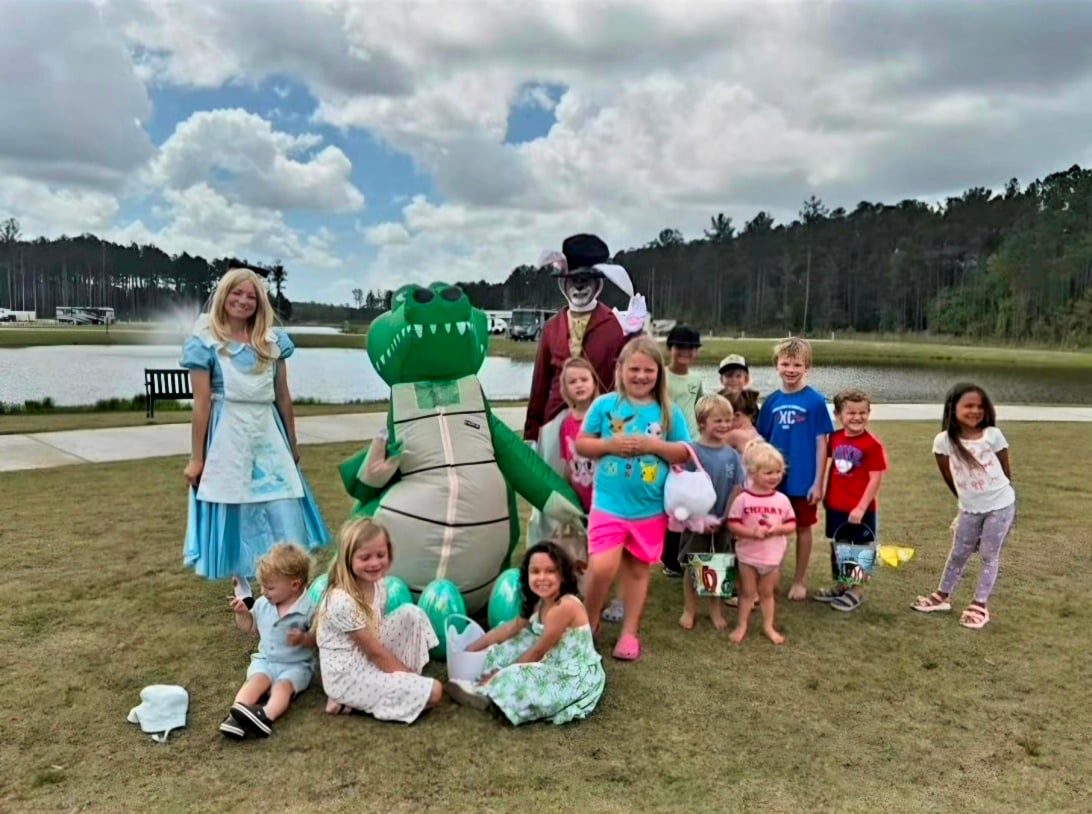 A group of children pose outdoors with adults in costumes, including a princess, an alligator, and a man in a top hat, near a pond and trees under a cloudy sky.
